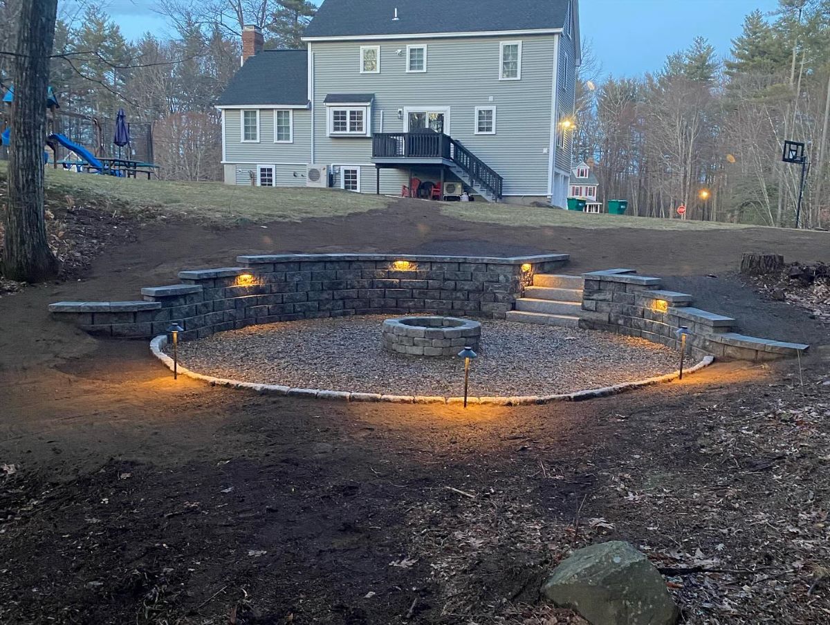 A circular outdoor fire pit area with built-in lighting and stone seating walls, located in a backyard with a large gray house in the background.