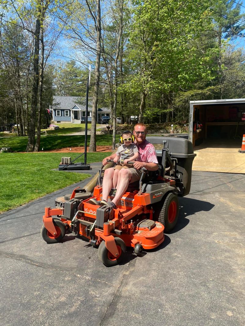 Man and child sitting on a large orange lawn mower in a driveway.