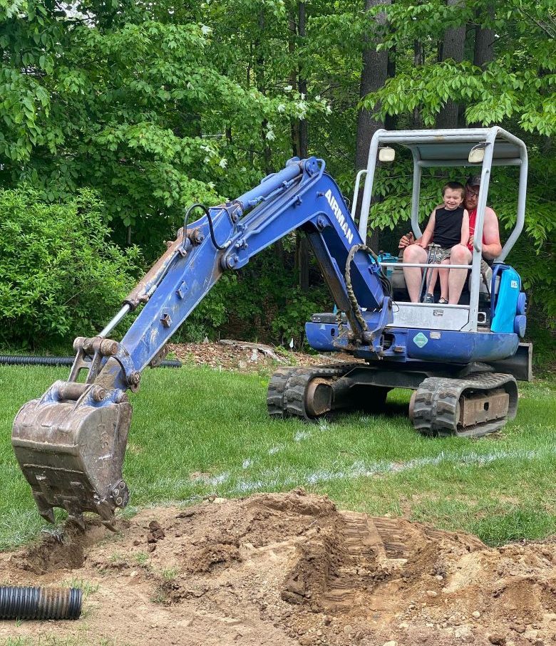 Two people operating a small blue excavator digging in a grassy yard.