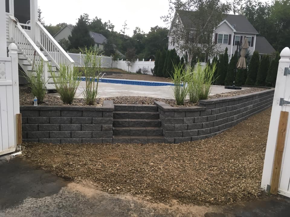 Elevated pool area surrounded by a dark stone retaining wall with stairs and ornamental grass plants, located in a fenced backyard.