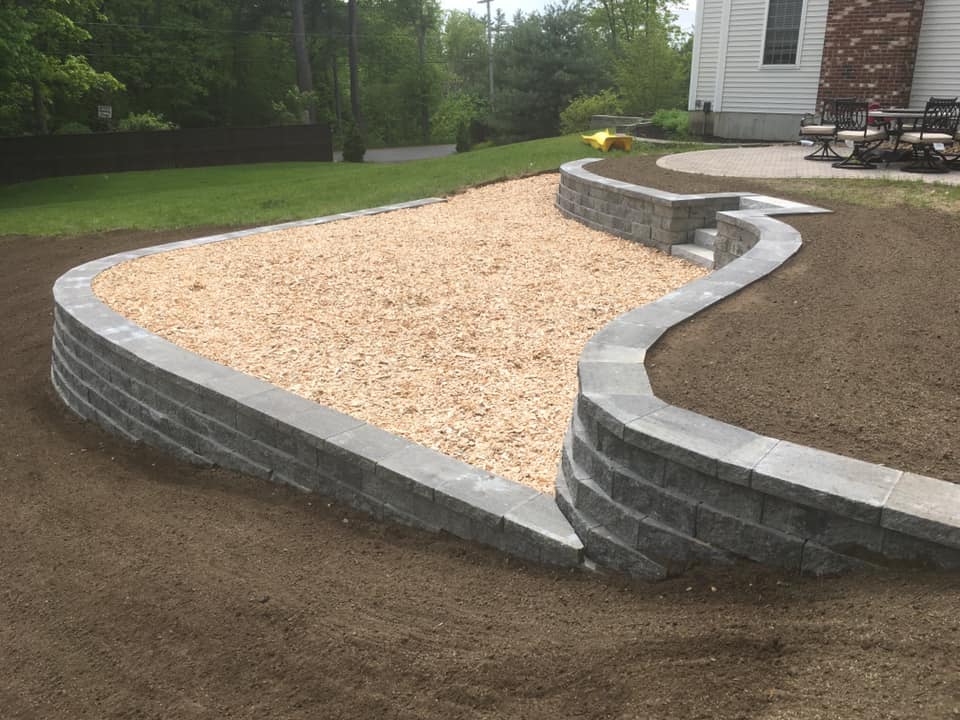 Curved retaining wall surrounding a raised area filled with light-colored gravel, located next to a house with bare soil around it, ready for planting or grass.