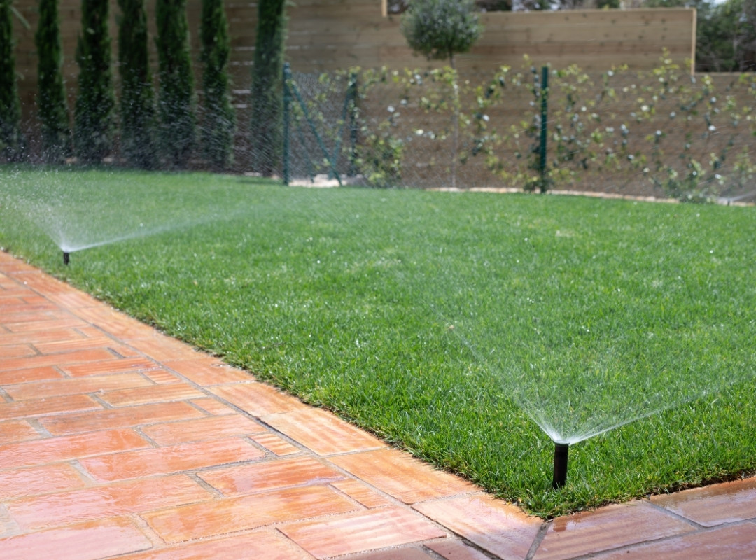 Lawn sprinklers watering a neatly maintained grassy area next to a tiled patio.
