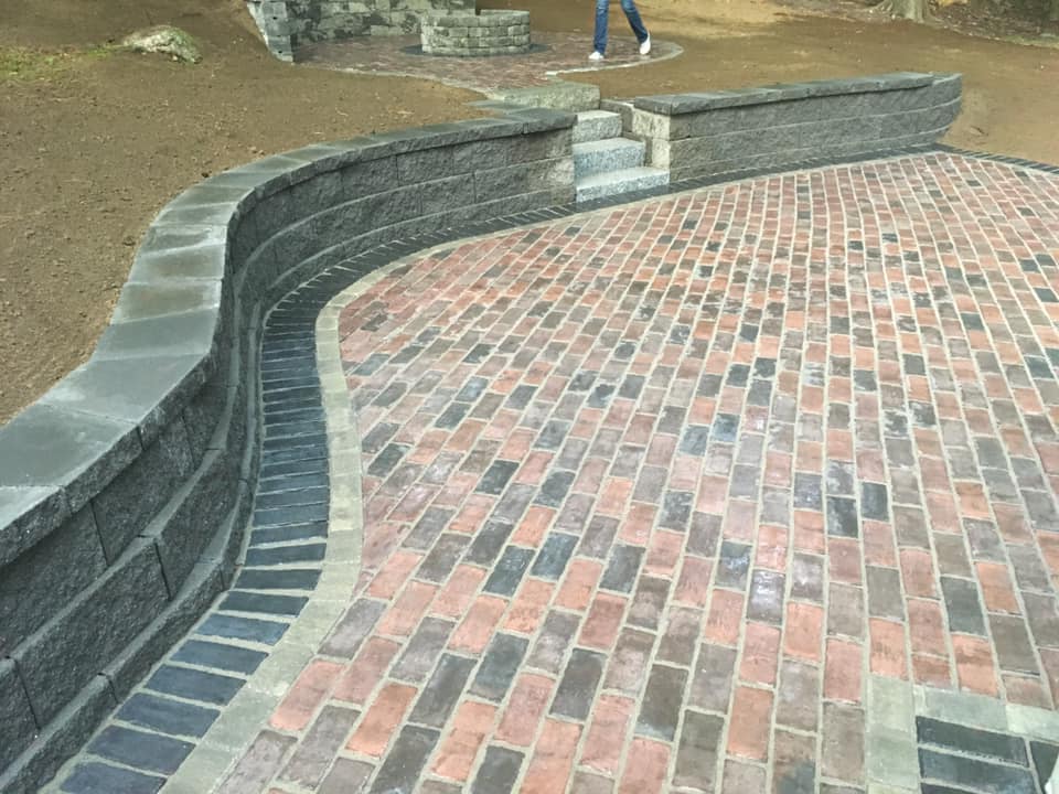 Brick paver patio with a curved retaining wall and small stone steps leading up to another tiered area.
