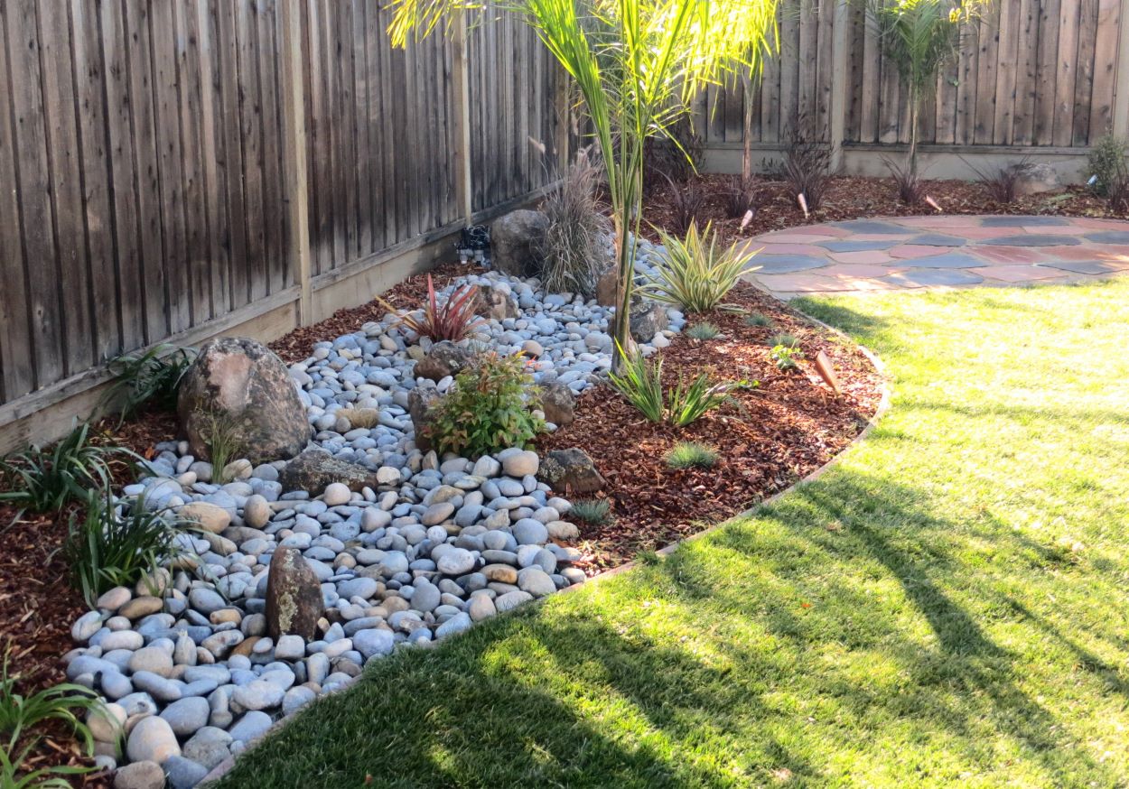 Backyard landscape with decorative rocks, mulch, and palm plants along a wooden fence.